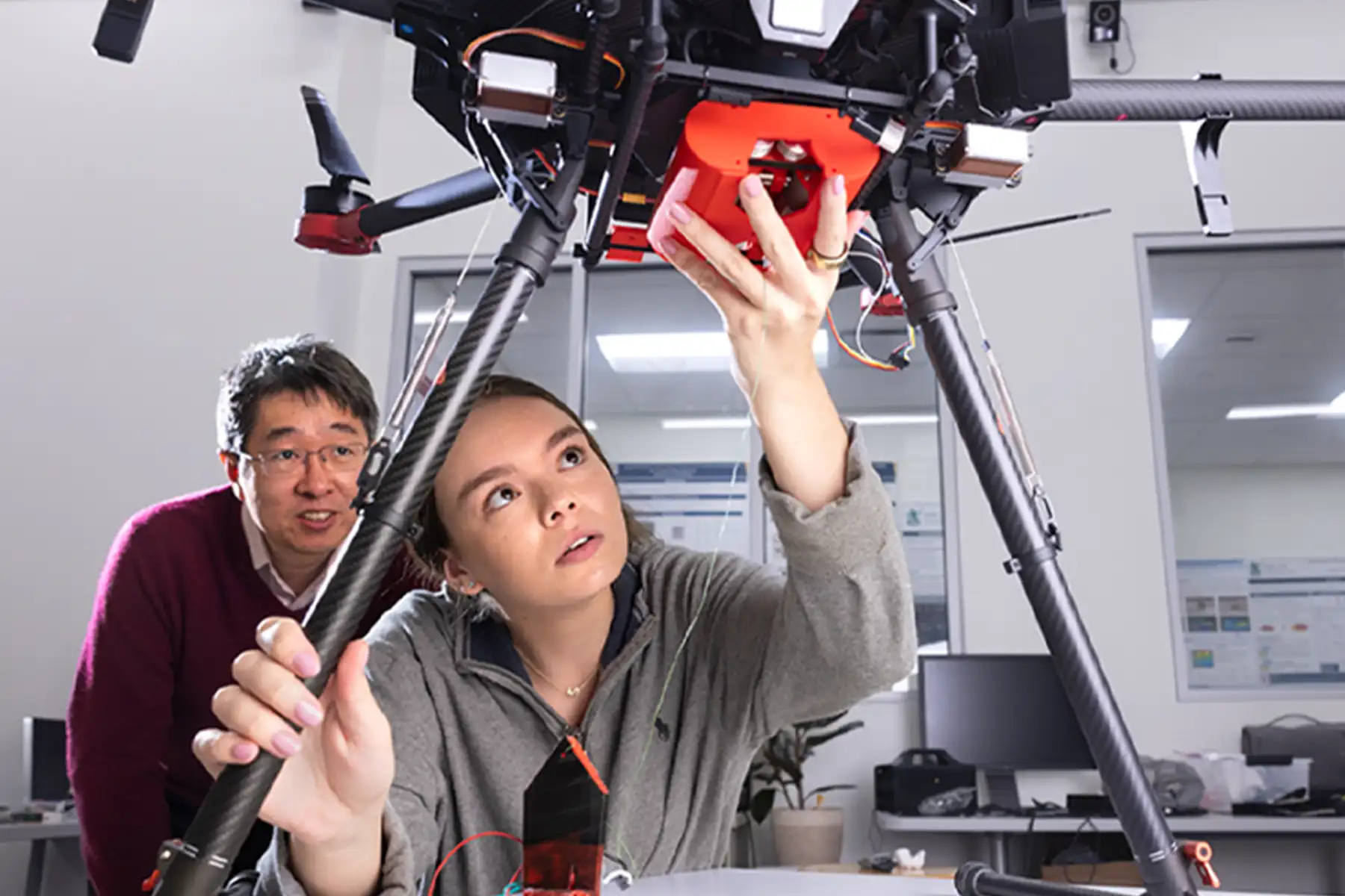 A female student looks up in wonder while working under a large drone, with a male professor watching her intently. 